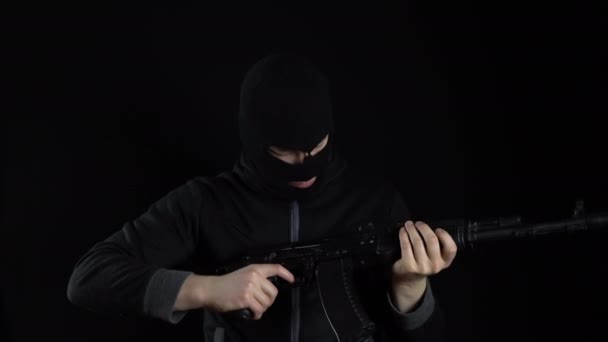 Un homme portant un masque de cagoule porte un fusil d'assaut AK-47. Le bandit charge la machine et se tient debout. Sur fond noir. — Video