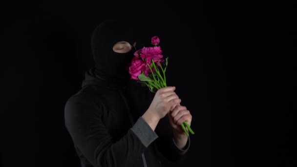 バラクラバマスクの男が花と一緒に立っている。バンディットは黒の背景にピンクの花の花束を嗅ぎます. — ストック動画