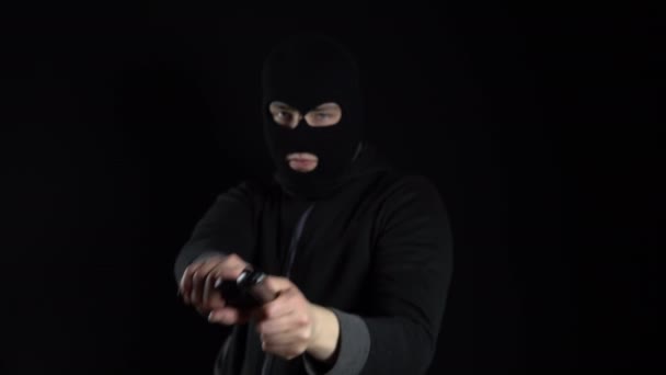 Чоловік у балаклавській масці стоїть з пістолетом. Бандит вказує на пістолет і стріляє в камеру. На чорному тлі . — стокове відео