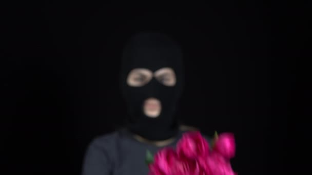 En kvinna i balaklavamask står med blommor. Bandit håller fram en bukett rosa blommor till kameran. På en svart bakgrund. — Stockvideo