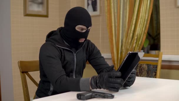 Um ladrão está a tentar invadir um tablet. Um bandido mascarado está sentado em uma casa e não pode invadir um tablet. Roubo de dados de um tablet. — Vídeo de Stock