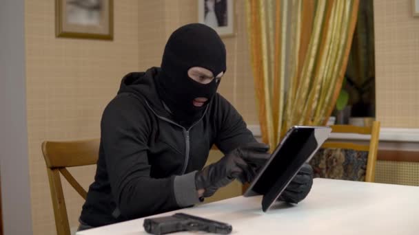 Um ladrão está a tentar invadir um tablet. Um bandido mascarado está sentado em uma casa tentando invadir um tablet e está ameaçando com uma arma. Roubo de dados de um tablet. — Vídeo de Stock