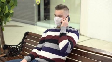 A young man in a medical mask sits on a bench in a shopping center and talks on the phone. The man is sneezing. The masked man protects himself from the epidemic of the Chinese virus 