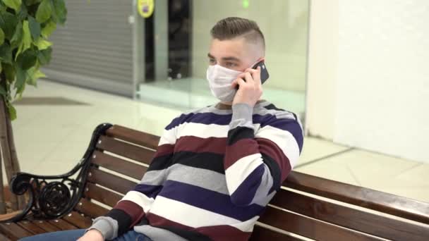 Un joven con una máscara médica se sienta en un banco en un centro comercial y habla por teléfono. El hombre estornuda. El hombre enmascarado se protege de la epidemia del virus chino "2019-nKoV " — Vídeos de Stock
