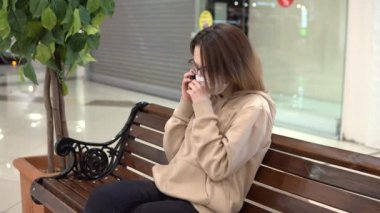 A young woman in a medical mask sits on a bench in a shopping center and talks on the phone. The woman is coughing. The masked woman protects herself from the epidemic of the Chinese virus 