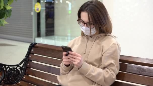 Μια νεαρή γυναίκα με ιατρική μάσκα κάθεται σε ένα παγκάκι σε ένα εμπορικό κέντρο και μιλάει στο τηλέφωνο. Η μασκοφόρος προστατεύει τον εαυτό της από την επιδημία του κινεζικού ιού "2019-nkov". — Αρχείο Βίντεο