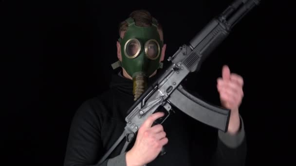 A man in a gas mask stands with a Russian AK-47 assault rifle. The bandit charges the machine with a machine. On a black background. — Stockvideo