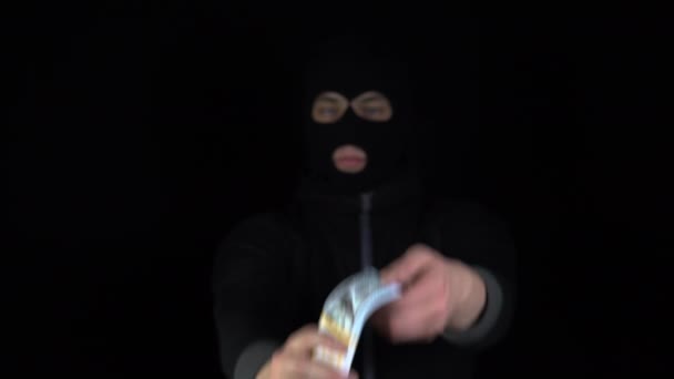 Un hombre con una máscara de pasamontañas sostiene un fajo de dólares. Bandido hojeando el dinero. Sobre un fondo negro. — Vídeo de stock