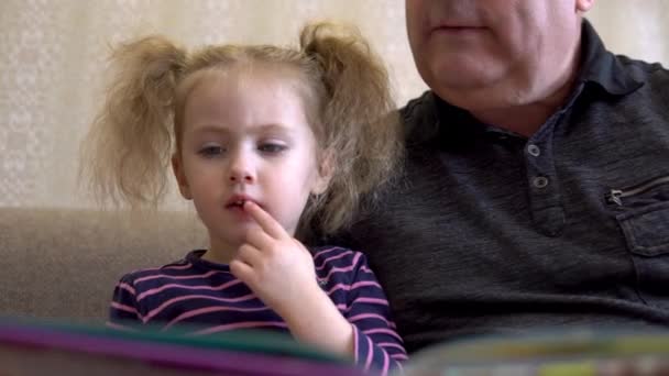 Sa petite-fille lit un livre avec son grand-père. La fille fronce les sourcils au livre et écoute attentivement Grand-père. Assis sur le canapé ensemble gros plan — Video