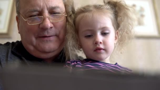 Petite-fille et grand-père sont assis avec une tablette. La fille fronce les sourcils à la tablette. Assis sur le canapé ensemble gros plan. — Video