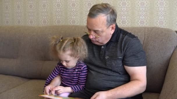 Дідусь читає книжку з дідусем. Дівчина насупилася на книжку і уважно вислуховує дідуся. Сидіння на дивані разом з "єднано. — стокове відео