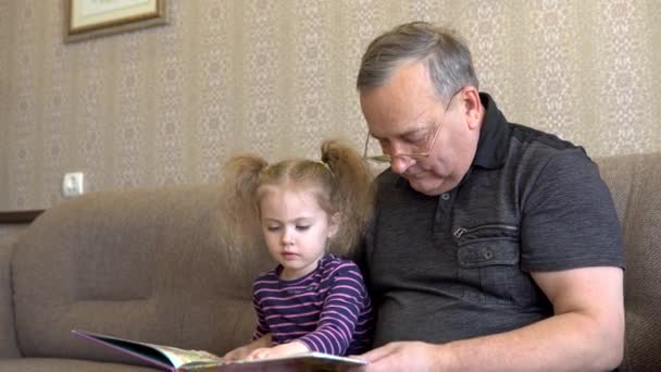 Wnuczka czyta książkę z dziadkiem. Dziewczyna marszczy brwi na książkę i uważnie słucha dziadka. Siedząc na kanapie razem zbliżenie — Wideo stockowe