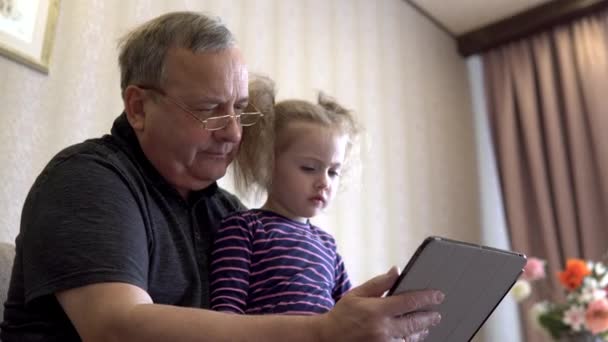 Enkelin und Großvater sitzen mit einem Tablet. Das Mädchen runzelt die Stirn angesichts der Tablette. Gemeinsam auf der Couch sitzen. — Stockvideo