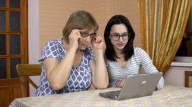 Daughter teaches mother how to use a laptop. A young woman shows her old mother where to click on a computer. The family is sitting in a comfortable room.