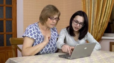 Kızı annesine dizüstü bilgisayar kullanmayı öğretiyor. Genç bir kadın yaşlı annesine bilgisayarın neresine tıklanacağını gösterir. Aile rahat bir odada oturuyor. Kadın kameraya bakıyor ve başparmağını gösteriyor.