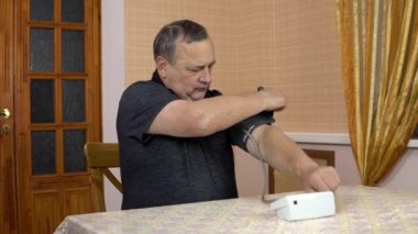 A man uses a device to measure heart pressure. An elderly man puts a blood pressure monitor on his hand and measures the pressure. A man sits in a comfortable room.