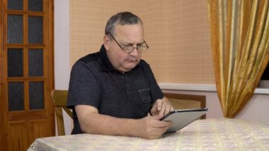A man uses a tablet for the first time. The old man does not understand how to use the tablet, pokes his finger, and then puts it off. A man in a comfortable room.