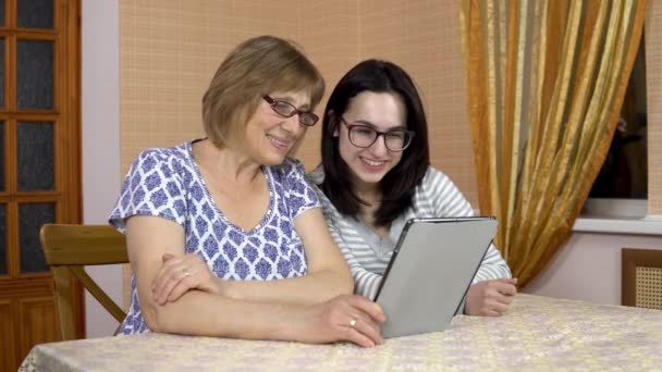 La hija y la Madre se comunican con los amigos a través del enlace de vídeo a través de la tableta. Una joven y una vieja madre se comunican y saludan con las manos, mirando una tableta. La familia está sentada en — Vídeo de stock