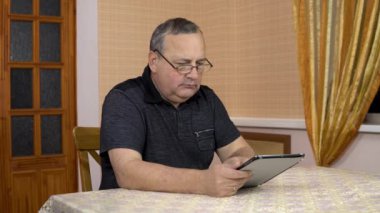 Serious man uses a tablet. Old man works with a tablet while sitting at the table. A man in a comfortable room.