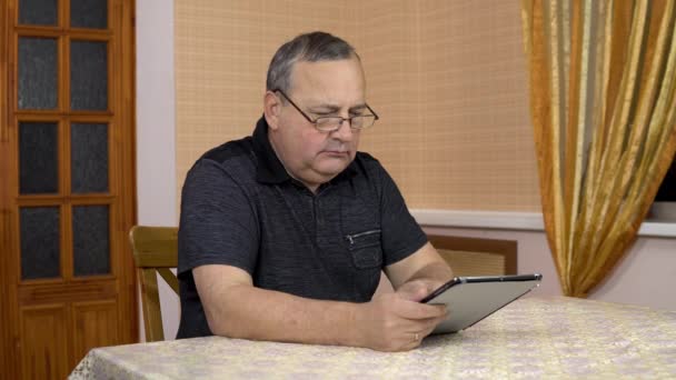 Serious man uses a tablet. Old man works with a tablet while sitting at the table. A man in a comfortable room. — Stock video