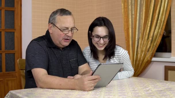 De dochter en haar vader communiceren met vrienden via videolink via de tablet. Een jonge vrouw en een oude man communiceren en zwaaien met hun handen, kijkend naar een tablet. De familie zit in een — Stockvideo