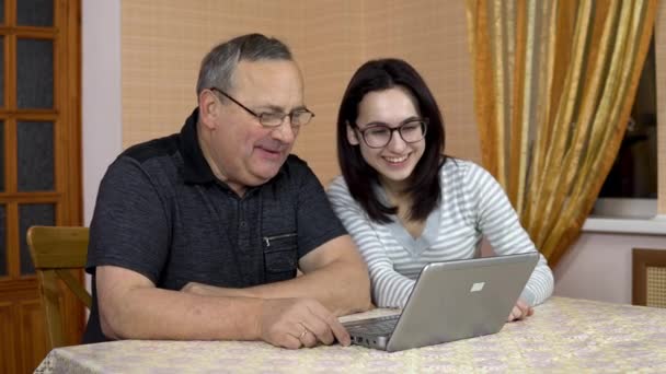 Uma filha e seu pai se comunicam com amigos através de comunicação por vídeo através de um laptop. Uma jovem e um velho pai se comunicam e acenam com as mãos enquanto olham para um laptop. A família é — Vídeo de Stock