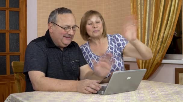 Mężczyzna i kobieta komunikują się z przyjaciółmi przez komunikację wideo przez laptopa. Starzec i starsza kobieta komunikują się i machają rękami patrząc w laptopa. Rodzina w nim siedzi. — Wideo stockowe