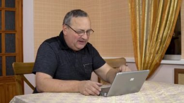 A man communicates with friends via video calling through a laptop. An old man communicates while looking at a laptop. A man in a comfortable room.
