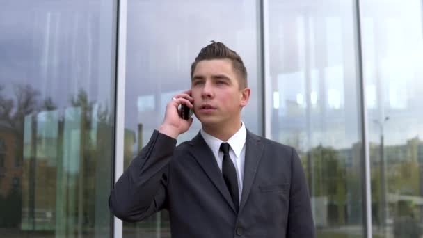 Un joven hombre de negocios con traje habla por teléfono. Un hombre serio se para frente a un centro de negocios de espejos. Movimiento lento — Vídeos de Stock