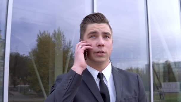 Un joven hombre de negocios con traje habla por teléfono. Hombre serio se para frente a un centro de negocios de espejos — Vídeo de stock