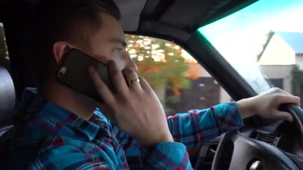 Un joven conduce un coche y habla por teléfono durante el día. Vista lateral del conductor — Vídeos de Stock