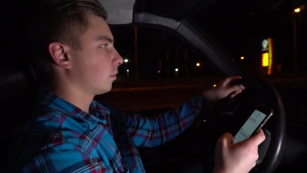 A young man drives a car and writes a message on the phone. Night time of the day. Driver side view — 비디오