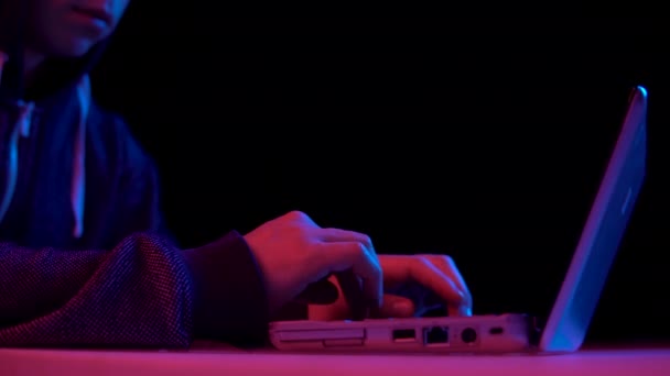 Young man in a hood with a laptop closeup. Hacker makes a hack through a laptop. Blue and red light falls on a man on a black background. — Wideo stockowe