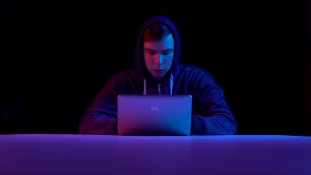 Young man in a hood with a laptop. Hacker makes a hack through a laptop. Blue and red light falls on a man on a black background. — ストック動画