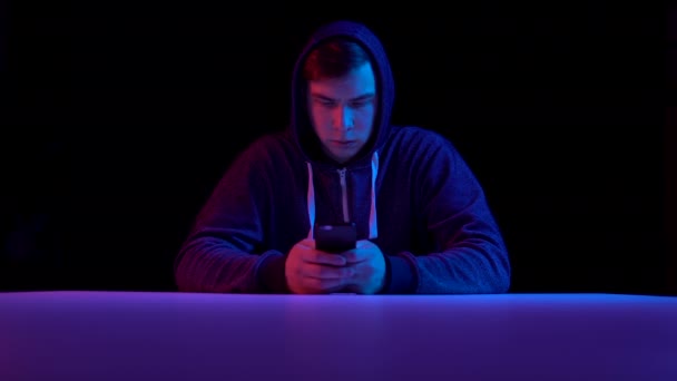 Young man in a hood with a smartphone. Hacker makes a hack through the phone. Blue and red light falls on a man on a black background. — 비디오