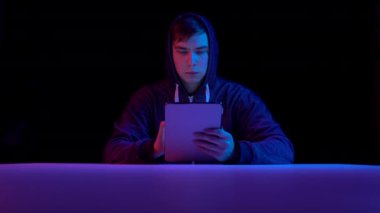 Young man in a hood with a tablet. Hacker makes a hack through the tablet. Blue and red light falls on a man on a black background.