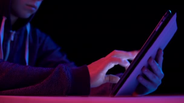 Young man in a hood with a tablet. Hacker makes a hack through the tablet. Blue and red light falls on a man on a black background. — Wideo stockowe