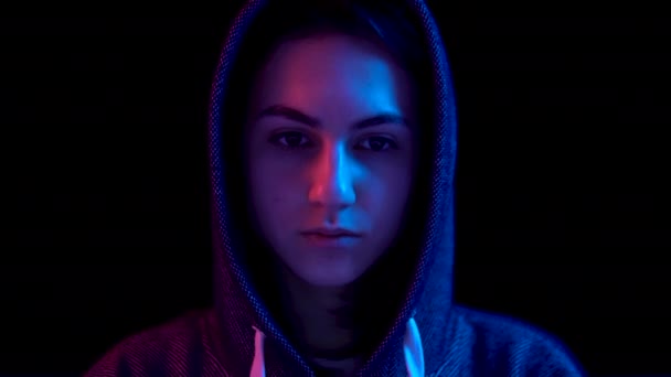 Young woman in a hood with a laptop. Hacker makes a hack through a laptop. Blue and red light falls on a woman on a black background. The camera goes down. — Αρχείο Βίντεο