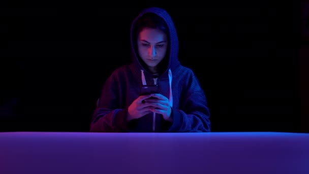 Young woman in a hood with a smartphone. Hacker makes a hack through the phone. Blue and red light falls on a woman on a black background. — Αρχείο Βίντεο