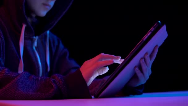 Young woman in a hood with a tablet. Hacker makes a hack through the tablet. Blue and red light falls on a woman on a black background. — Wideo stockowe