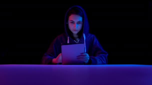 Young woman in a hood with a tablet. Hacker makes a hack through the tablet. Blue and red light falls on a woman on a black background. — Stok video