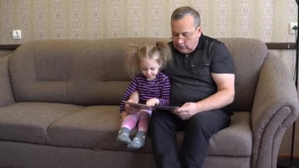 Sa petite-fille lit un livre avec son grand-père. La fille fronce les sourcils au livre et écoute attentivement Grand-père. Assis sur le canapé ensemble. — Video