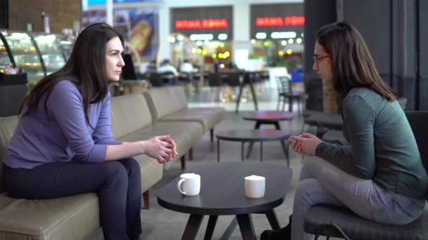 Le giovani donne sono sedute in un caffè a chiacchierare. Mamma e figlia si siedono l'uno contro l'altro, chiacchierando e bevendo caffè . — Video Stock