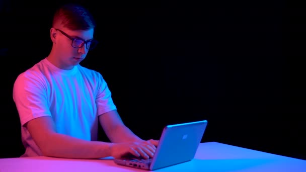 Un joven con un portátil. Un hombre usa un portátil. Luz azul y roja cae sobre un hombre sobre un fondo negro . — Vídeos de Stock