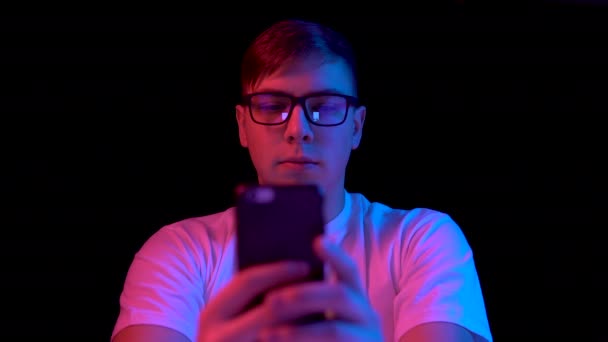 Young man with a smartphone. A man is using a telephone. Blue and red light falls on a man on a black background. — ストック動画