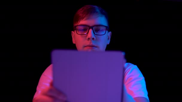 Young man with a tablet. A man is using a tablet. Blue and red light falls on a man on a black background. — Stok video