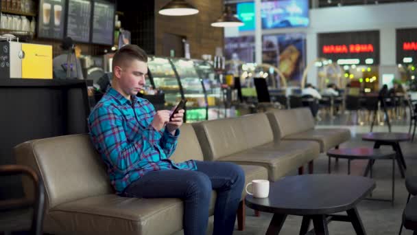 Un joven está sentado en un café con un teléfono. El hombre está en correspondencia en un teléfono inteligente y bebe café. Primer plano . — Vídeos de Stock