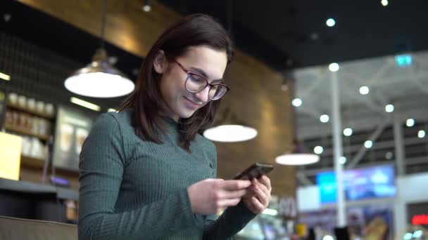 Una giovane donna è seduta in un caffè con un telefono. La ragazza è in corrispondenza su uno smartphone e beve caffè. Primo piano . — Video Stock