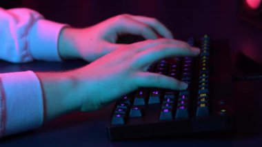 A young man is typing on a computer keyboard. Hands close up. Blue and red light falls on the hands.