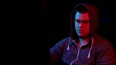 Genç bir adam bilgisayarın başında oturuyor. Kapüşonlu adam monitöre bakıyor..